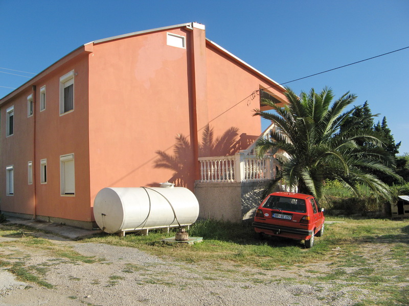 Two-story pink house in Bar, Montenegro: facade with a balustrade, a palm tree, and a water tank on the property.