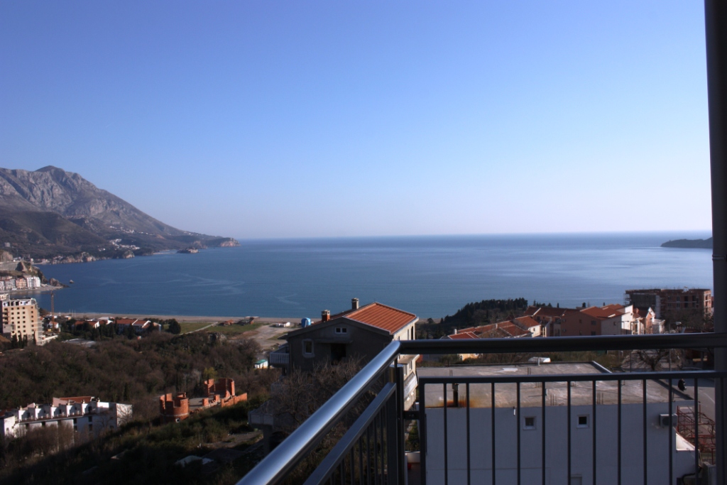 Panoramic view of the sea and mountains from the apartment balcony in Bečići, Montenegro, with a metal terrace railing.