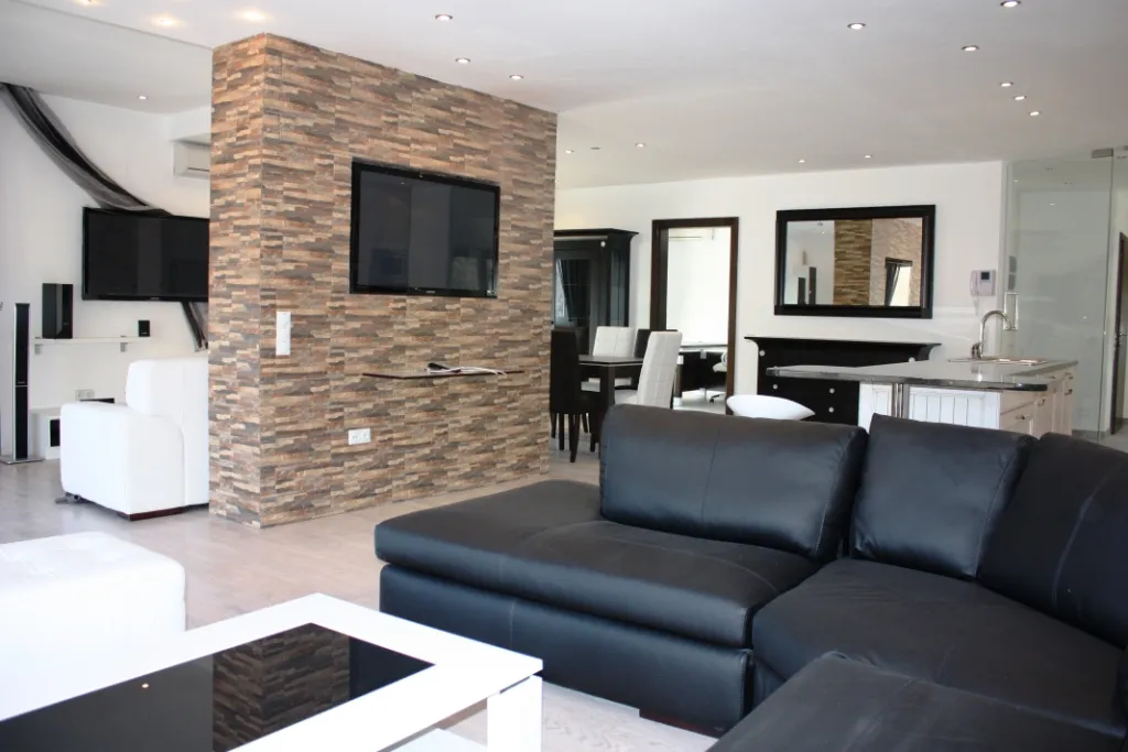 Modern living room with a black leather sofa, stone TV wall, white furniture, and open plan layout.