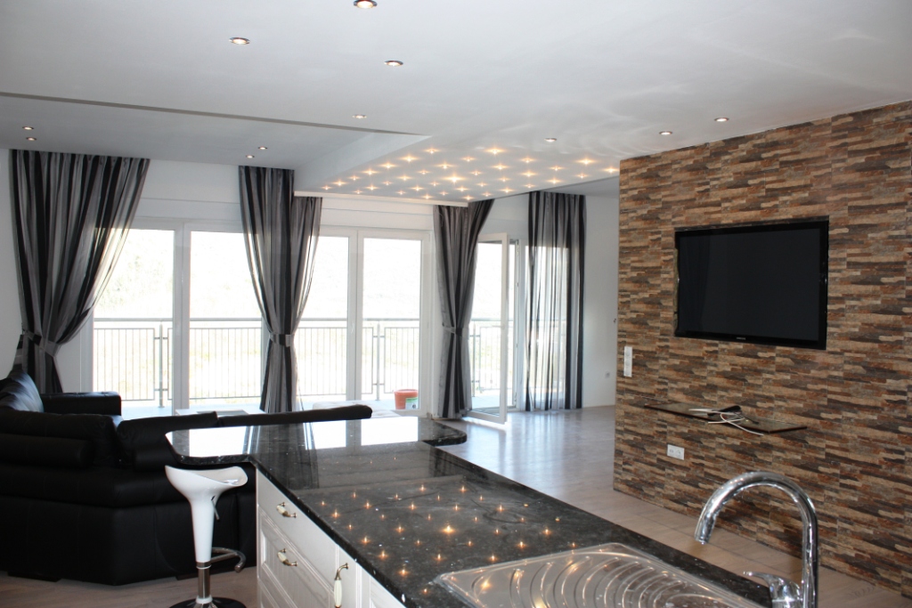 Modern living room with kitchen in an apartment in Bečići, Montenegro: stone accent wall, TV, panoramic windows, and a bar counter.