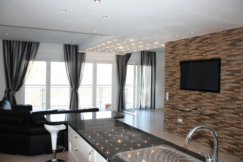 Modern open-plan living room with kitchen island, black sofa, TV on stone wall, and starry ceiling lights.