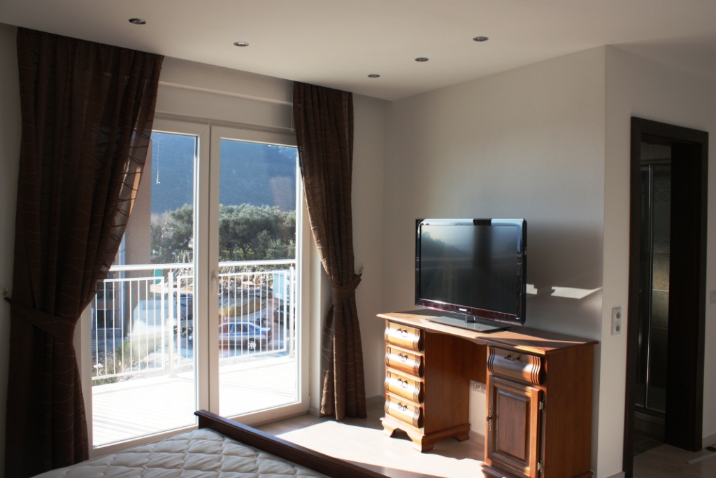 Bedroom in an apartment in Bečići, Montenegro: wooden dresser with TV, panoramic window with access to the balcony, and mountain view.