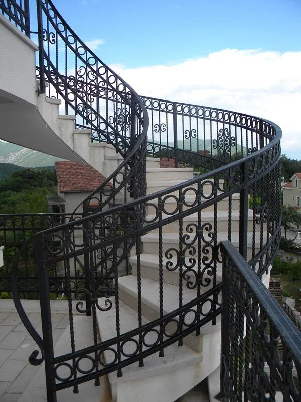 Spiral outdoor staircase with ornate wrought iron railing against mountains and sky.