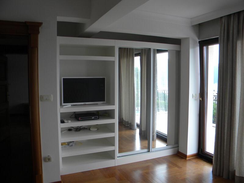 Living room with white built-in shelving unit holding a TV and mirrored wardrobe doors near a window.
