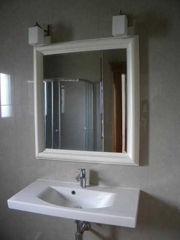 White sink with a white-framed mirror and wall lights above it on a grey tiled wall.
