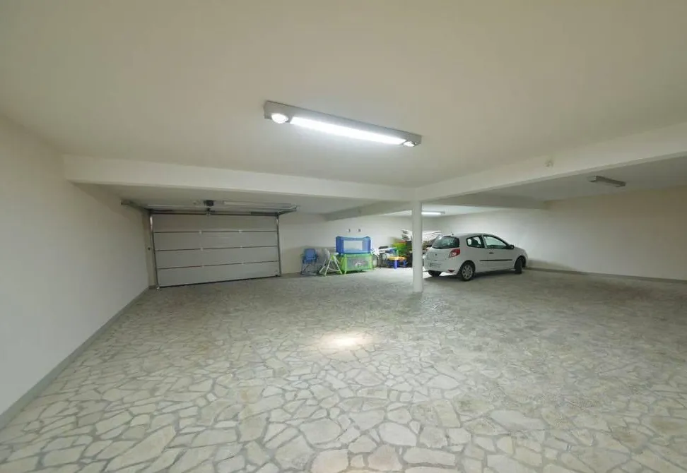 Garage containing a white car, sectional door, and stone floor. Storage items and ceiling light are visible in the spacious room.