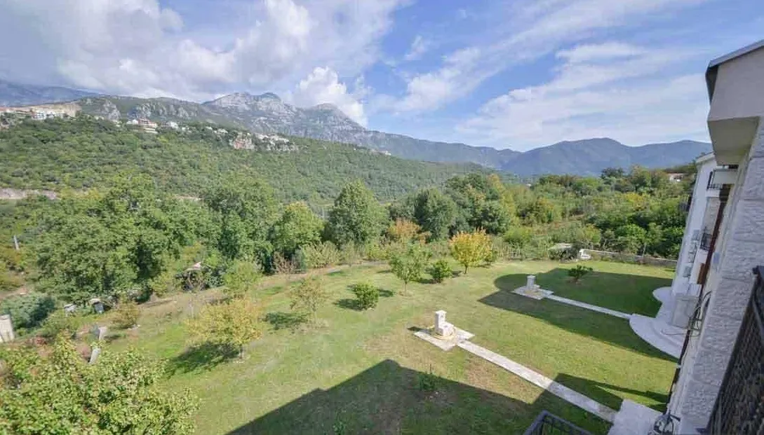 View of a green garden and mountains from a building balcony under a cloudy sky.