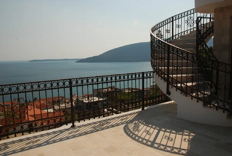 Terrace with ornate iron railing and curved staircase overlooking the sea and hills.