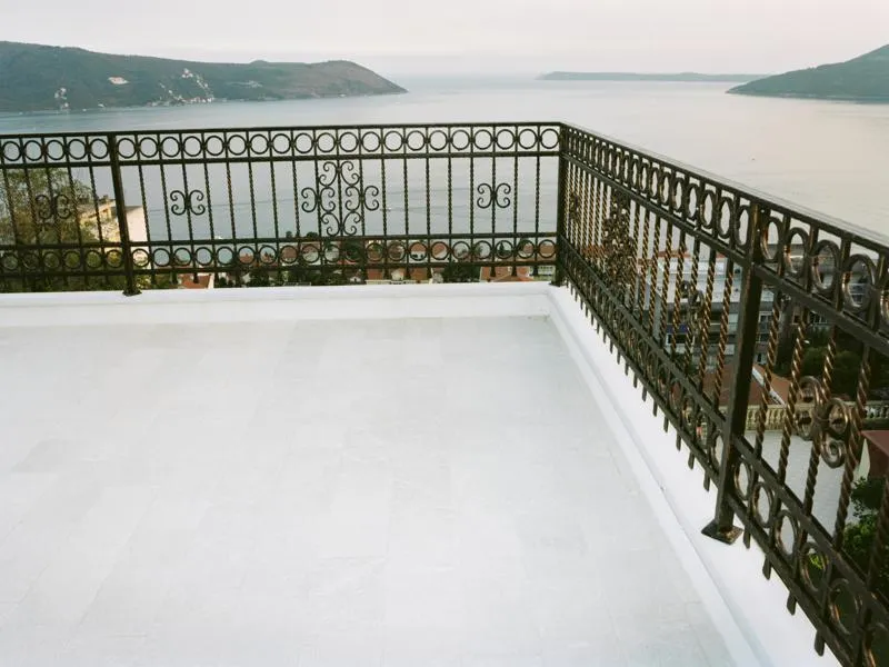 Balcony with white tiled flooring and ornate wrought iron railing overlooking the sea and hills.