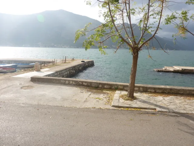 Street scene with a tree, stone embankment, and boats against a backdrop of water and mountains.