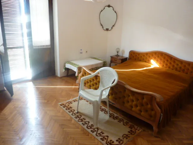 Bedroom featuring a wooden bed with a gold cover, white plastic chair, patterned rug on parquet flooring.