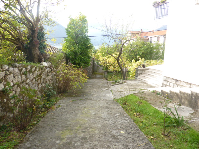 Concrete path to the house in Kumbor, Montenegro, with a stone fence, garden, and sea view surrounded by greenery.