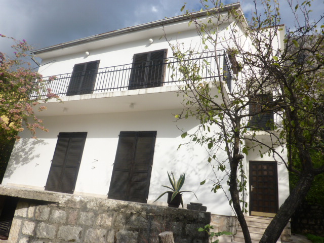 Two-story white house with a balcony and dark shutters in Kumbor, Montenegro, with mountains and a garden in the background.