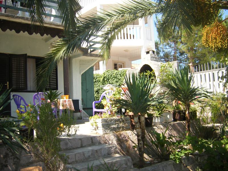 Cozy house terrace in Tivat, Montenegro, with garden furniture, palm trees, and a stone-clad facade surrounded by greenery.