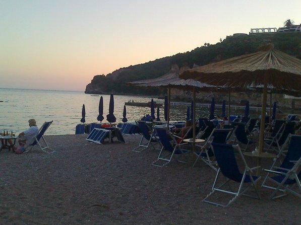 View of the beach with sun loungers and straw umbrellas by the sea in Budva, Montenegro, against a scenic rocky coastline.