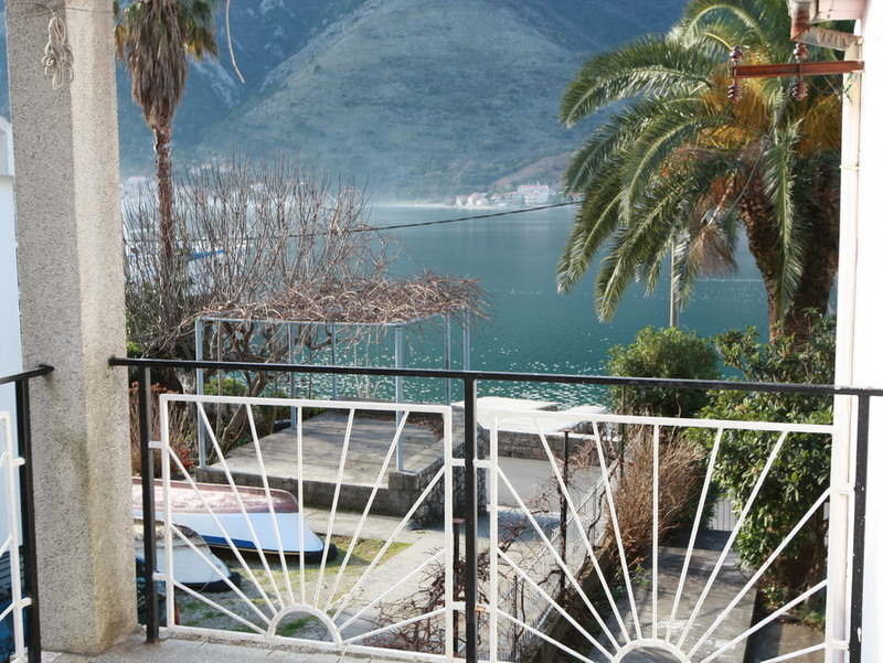 View from a balcony of a house in Dobrota, Montenegro: panoramic view of the Bay of Kotor, mountains, a palm tree, and boats on the shore.