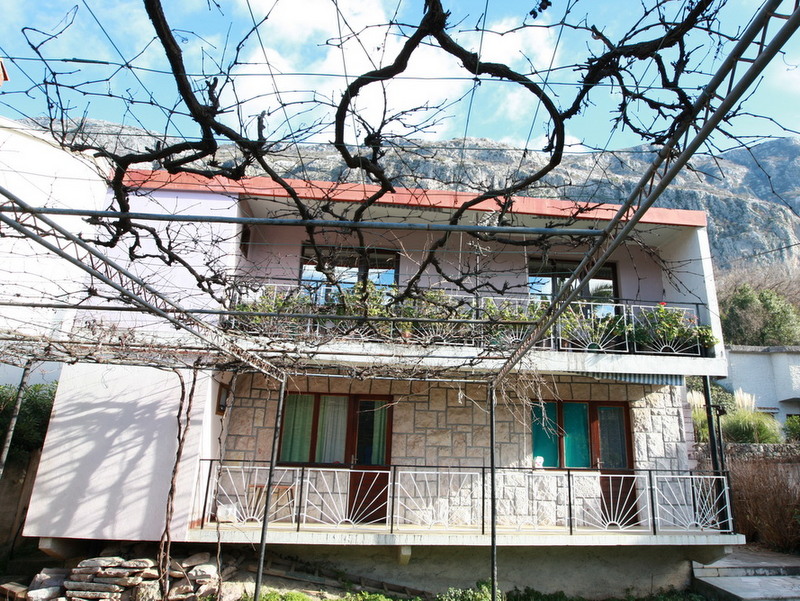 Two-story house in Dobrota, Montenegro, featuring stone finishes, terraces, and a grapevine canopy against a mountain backdrop.