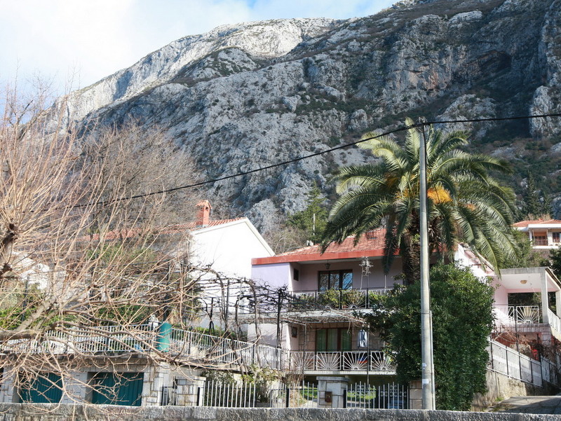 Two-story house with a terrace and a palm tree against the backdrop of mountains in Dobrota, Montenegro. View of a private house with a fence.