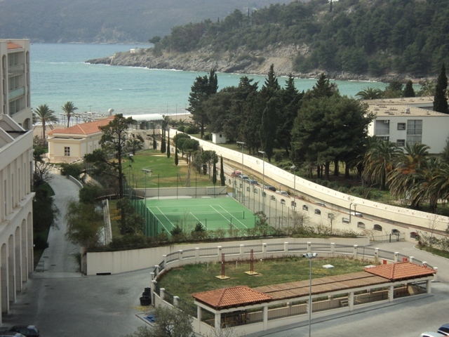 View of the tennis court, park area, and sea from an apartment in Bečići, Montenegro. Residential complex with amenities.