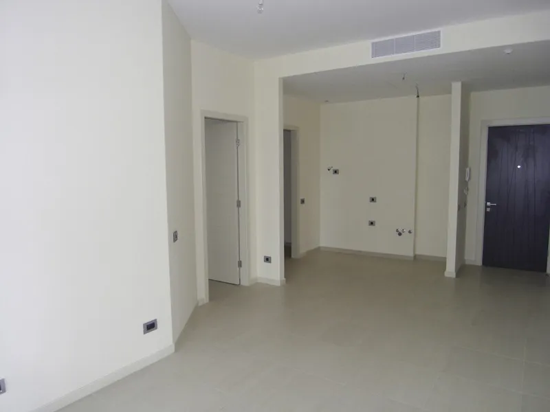 Empty room with light walls, tiled floor, and dark entrance door on the right. Visible electrical outlets and ceiling vent.
