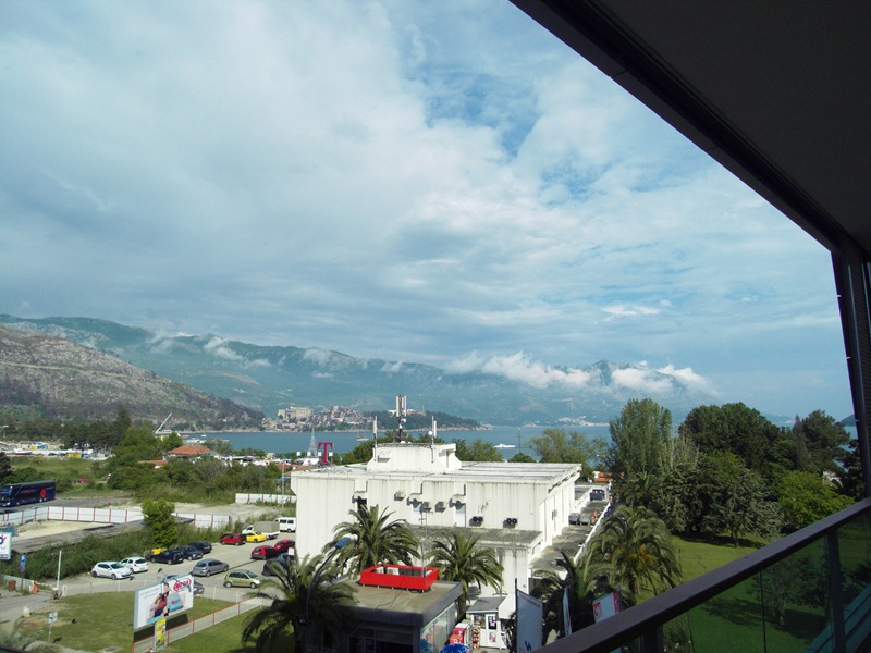 View from the apartment balcony in Budva, Montenegro: a panorama of mountains, the sea, and city infrastructure with palm trees.