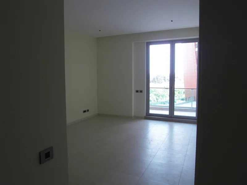 Empty room with tiled flooring and glass door leading to a balcony, visible electrical outlets on plain walls.