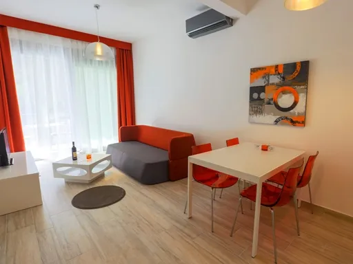 Living room featuring an orange sofa, white dining table, red transparent chairs, and abstract wall art.