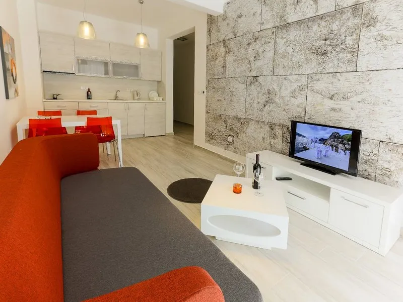 Living room with orange-grey sofa, white coffee table, TV on a textured stone wall, and kitchen in background.