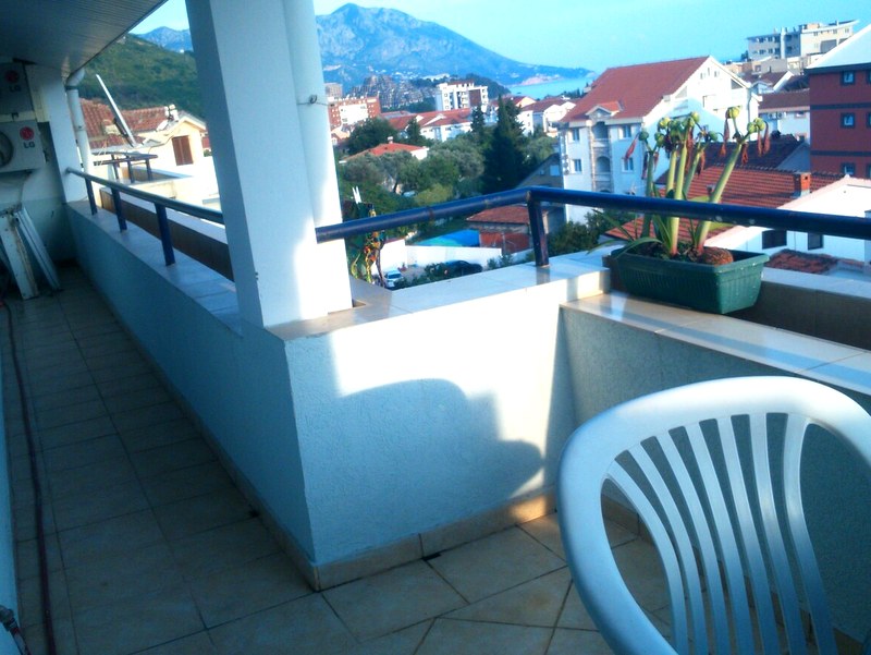 Spacious apartment balcony in Budva, Montenegro, with mountain, city, and sea views, plastic furniture and flowers.
