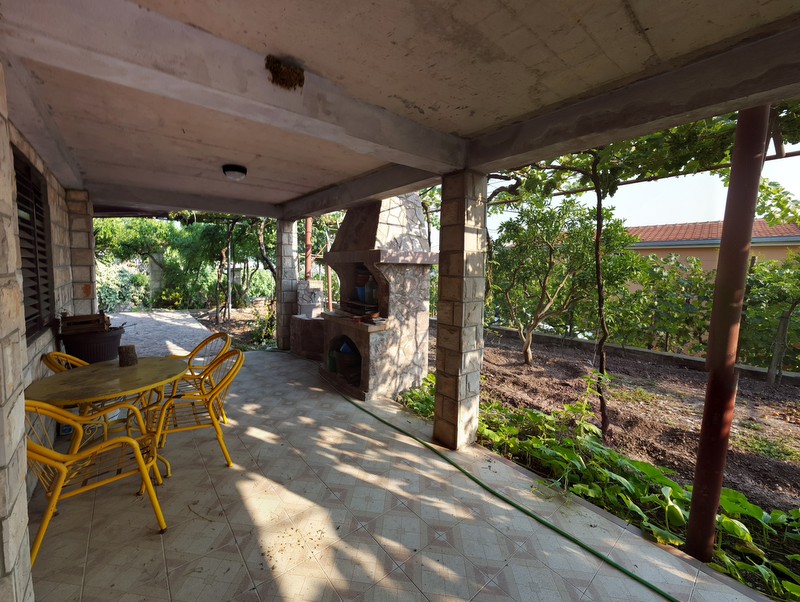 Covered terrace with stone barbecue and yellow furniture in a house in Shushan, Montenegro, overlooking a green garden and vineyard.