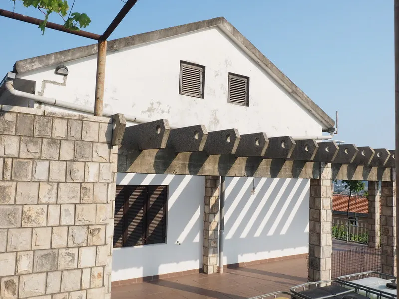 Exterior of a white house with a stone wall and concrete pergola under a clear sky.