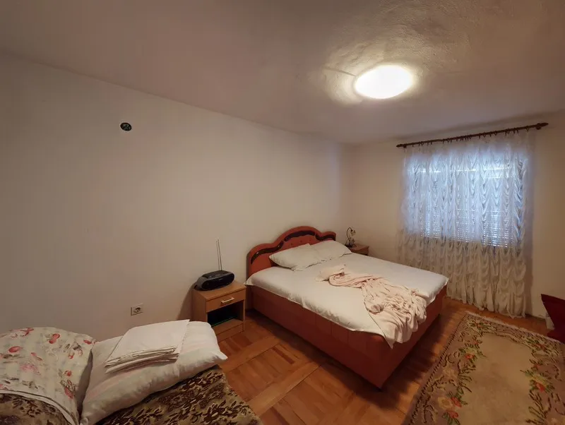 Bedroom featuring a wooden bed with white linens, a nightstand, sheer curtains, and a patterned rug on wood flooring.