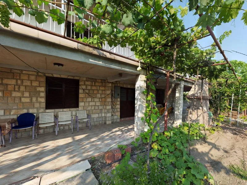 House exterior with stone wall, tiled terrace shaded by a grapevine pergola, and plastic chairs next to a garden.