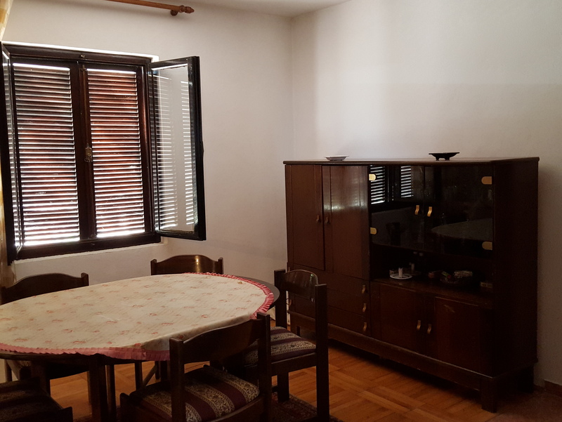 Living room in a house in Shushan, Montenegro: dining area with a wooden table, chairs, and a display cabinet, window with blinds.