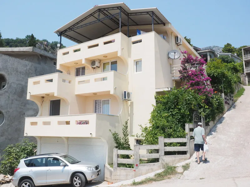Exterior view of a multi-story residential building with balconies, a parked car, and a man walking nearby.