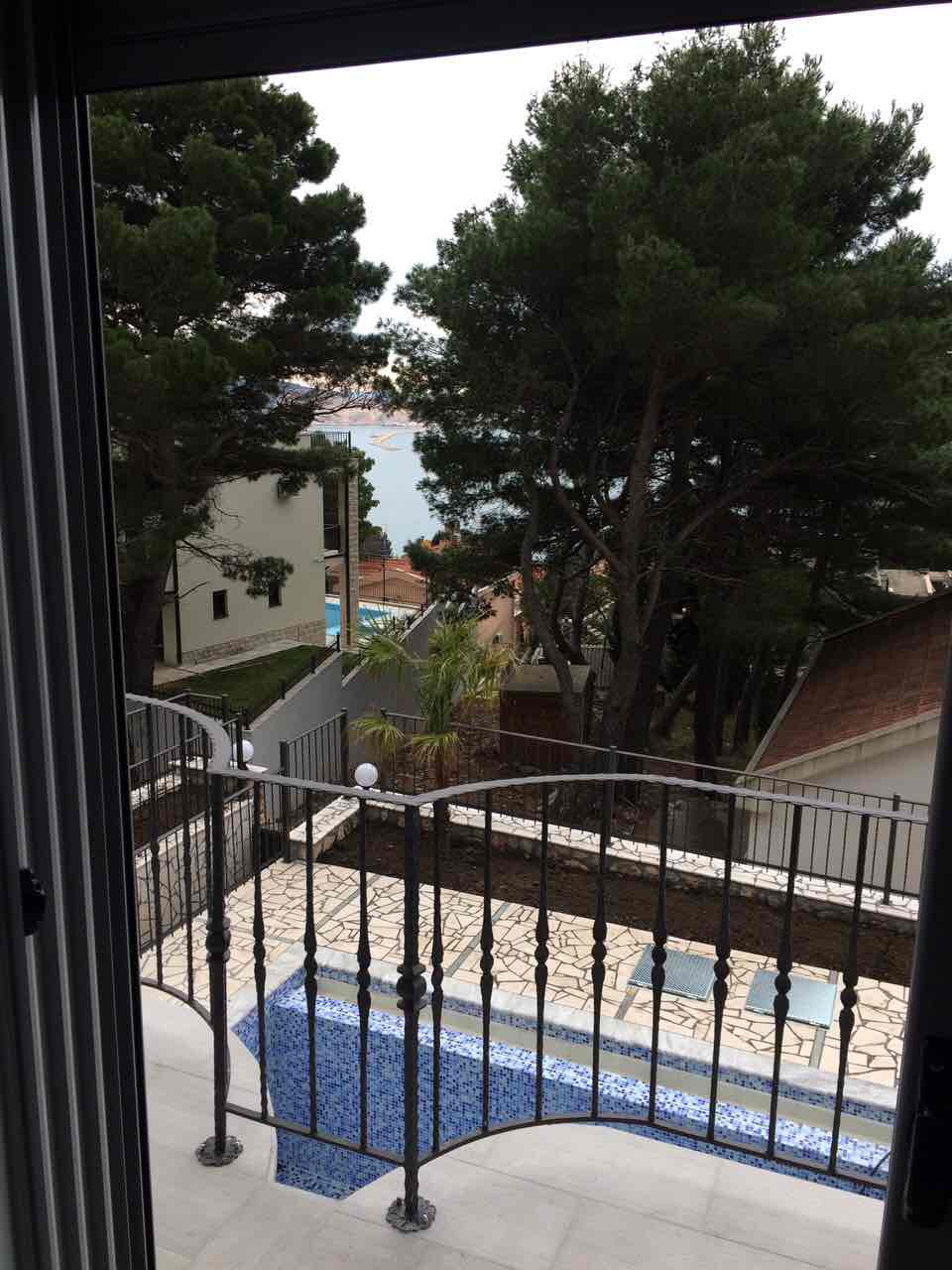 View from the balcony of a house in Shushan, Montenegro: a mosaic pool, terrace, conifer trees, and a sea view in the background.
