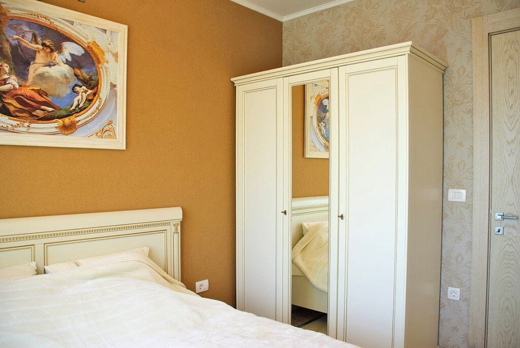 Bright bedroom with classic furniture and a painting in an apartment in Kostanica, Montenegro: a white bed and a wardrobe with a mirror.