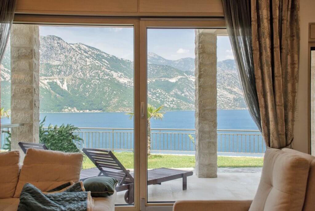 View from the living room of the sea and mountains in an apartment in Kostanica, Montenegro: panoramic windows, a terrace with sun loungers, and stone columns.
