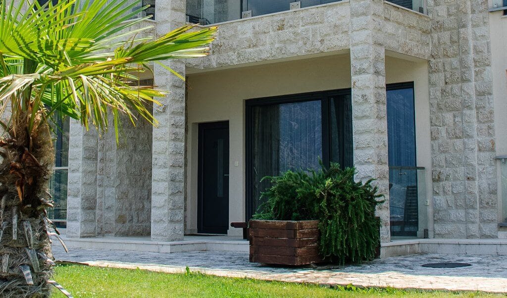 Facade of a modern apartment in Kostanica, Montenegro: stone cladding, panoramic windows, and a terrace with a palm tree.