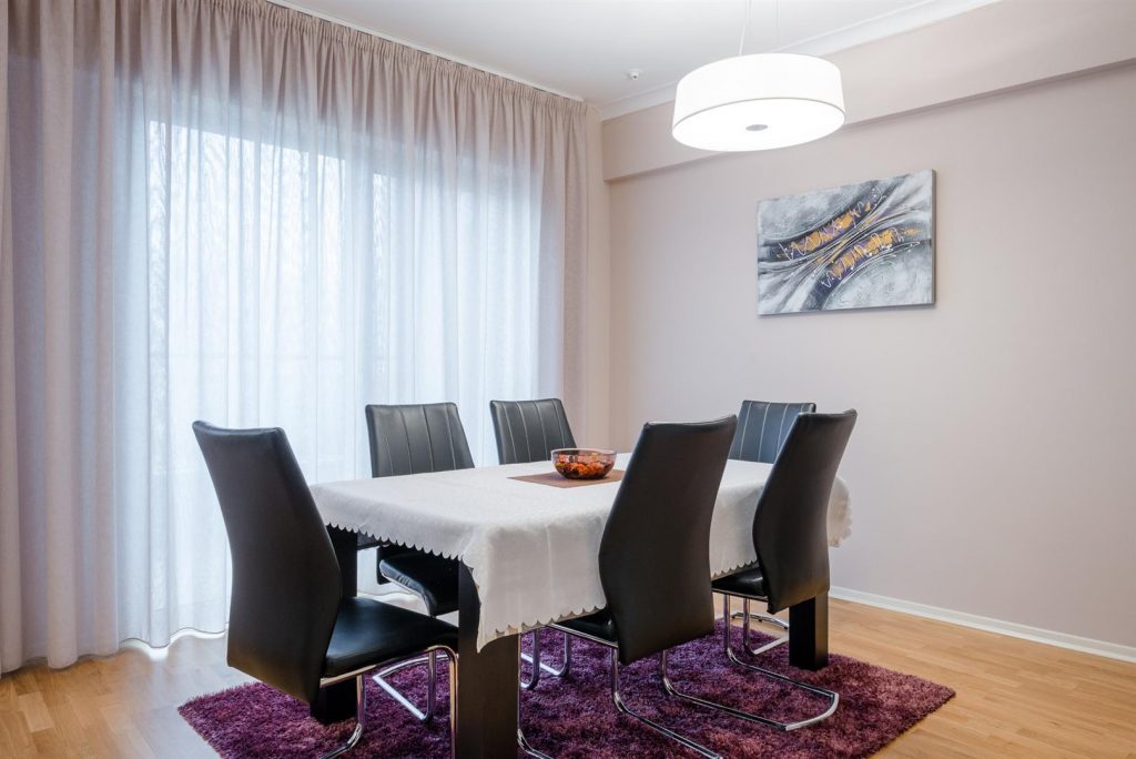 Dining area in an apartment in Bečići, Montenegro: table, black chairs, light curtains, and a painting in the living room interior.