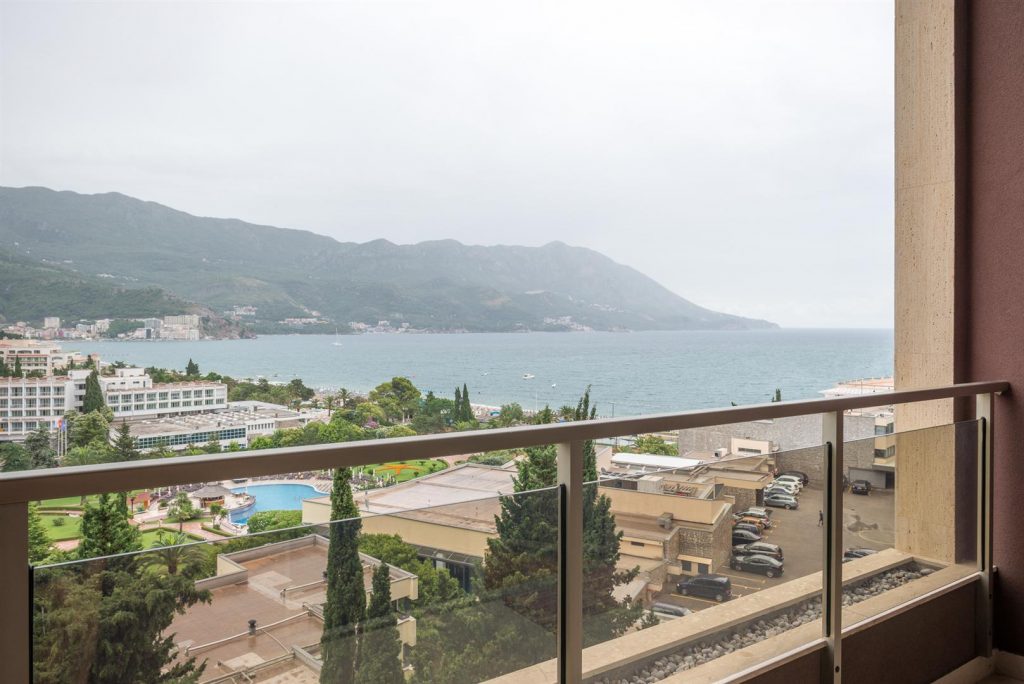 View from the apartment balcony in Bečići, Montenegro: panoramic sea and mountain views, hotel with a pool, glass railing.