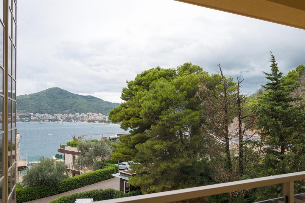 View from the apartment balcony in Bečići, Montenegro: a panorama of the sea, mountains, and green conifer trees near the building.