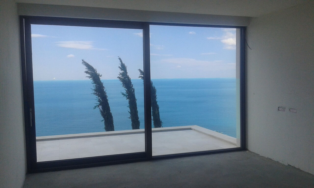 Panoramic window in the villa living room in Reževići, Montenegro, overlooking the sea and a terrace with cypress trees.