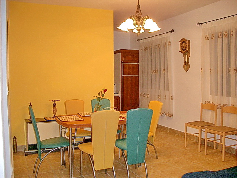 Living room interior with dining area in a hotel in Budva, Montenegro: yellow wall, wooden table, and colorful chairs.