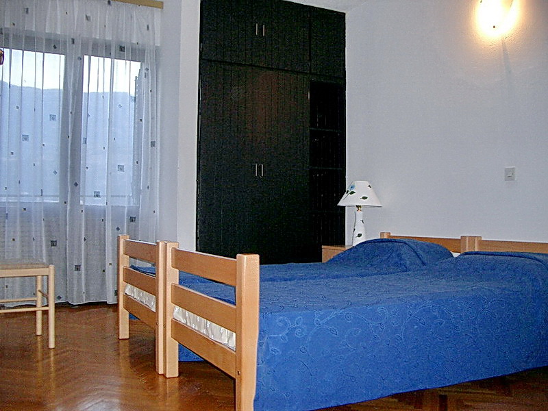 Bedroom with two beds, a dark wardrobe, and parquet flooring in a hotel in Budva, Montenegro. View of the balcony with sheer curtains.