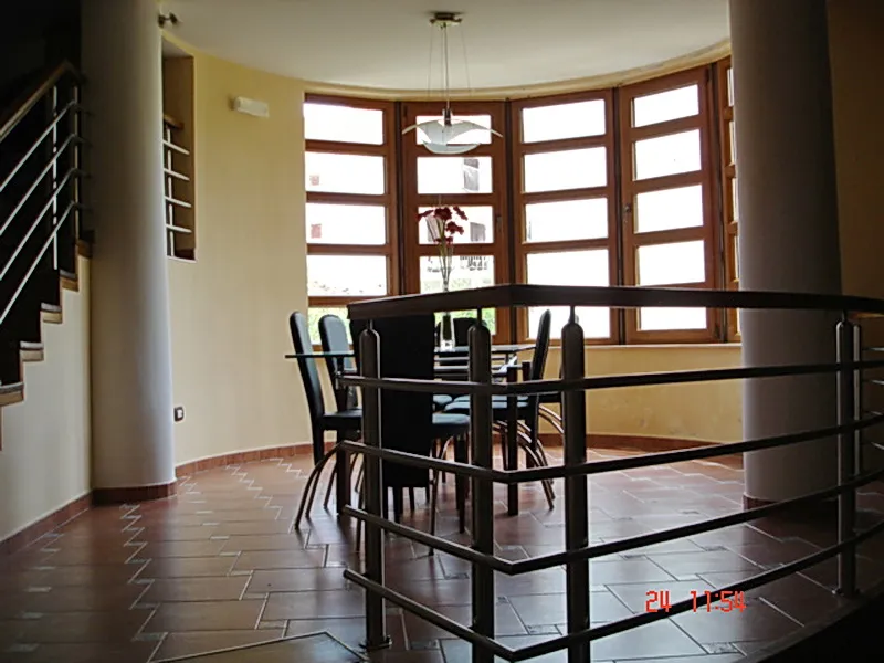 Dining area with black table and chairs near large windows, staircase visible on left, metal railing in foreground.