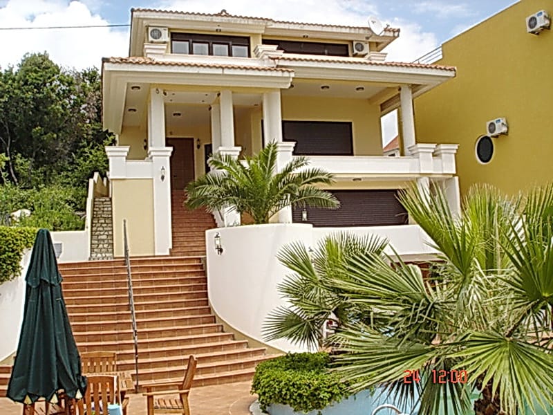 Two-story villa in Utjeha, Montenegro: facade with a terrace, wide staircase, palm trees, and a pool on the property.