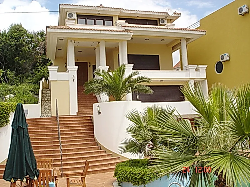 Two-story house exterior with terracotta stairs, white columns, palm trees, and a closed green umbrella in the foreground.