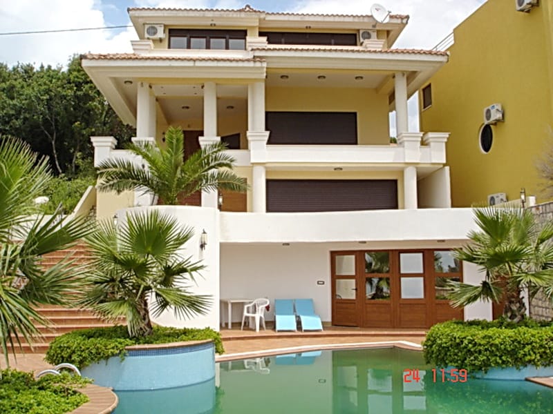Three-story villa with a pool, terraces, and palm trees in Utjeha, Montenegro. Facade with columns and a lounge area by the water.