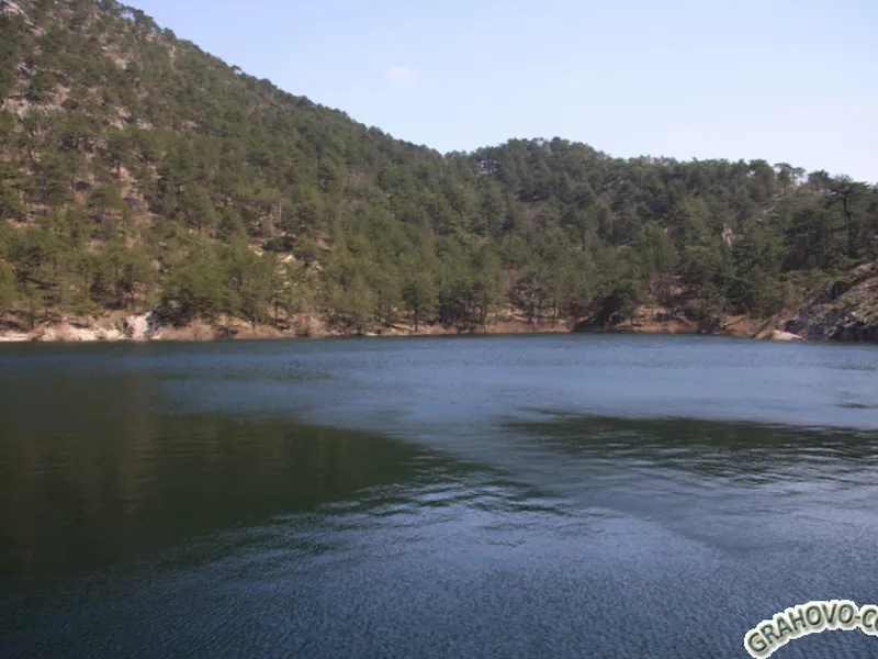 Natural landscape showing a lake or reservoir with a forested hill in the background under a clear sky.